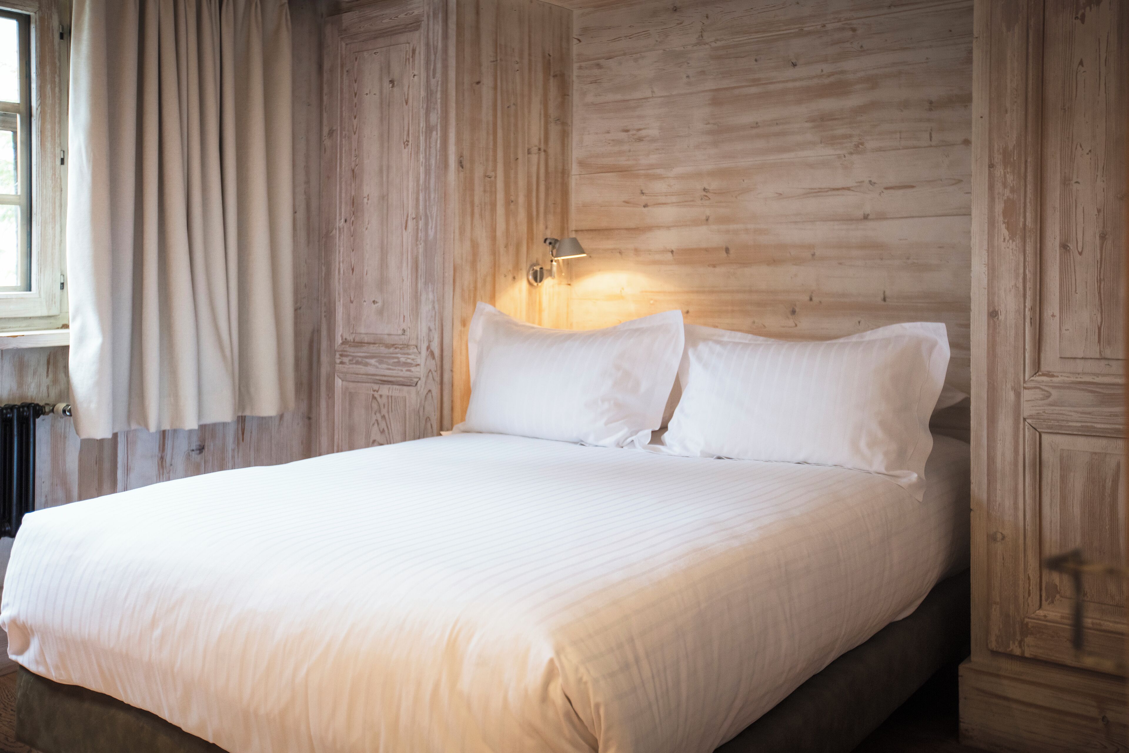 A neatly made bed with white linens in a room with wooden paneling and a window with curtains.