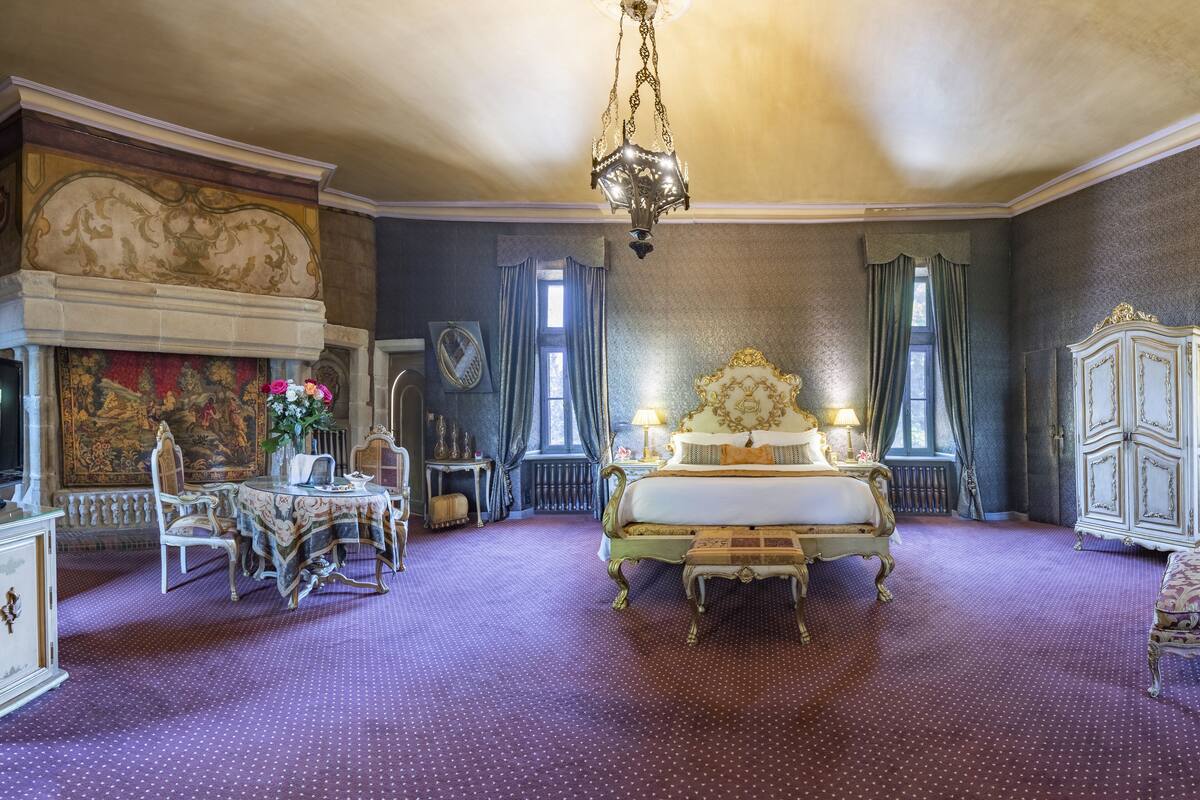 Una habitación amplia con una cama grande, un candelabro, una chimenea y un comedor con sillas y una mesa.