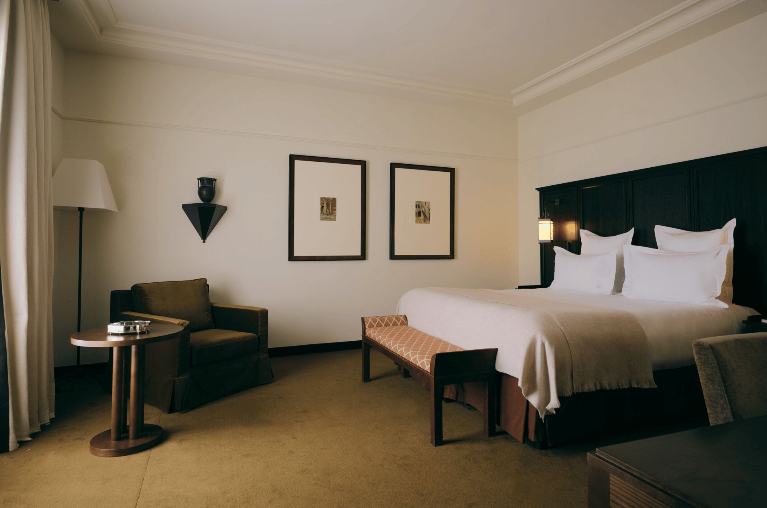 Habitación de hotel con cama, una silla, una mesita y dos cuadros en la pared.