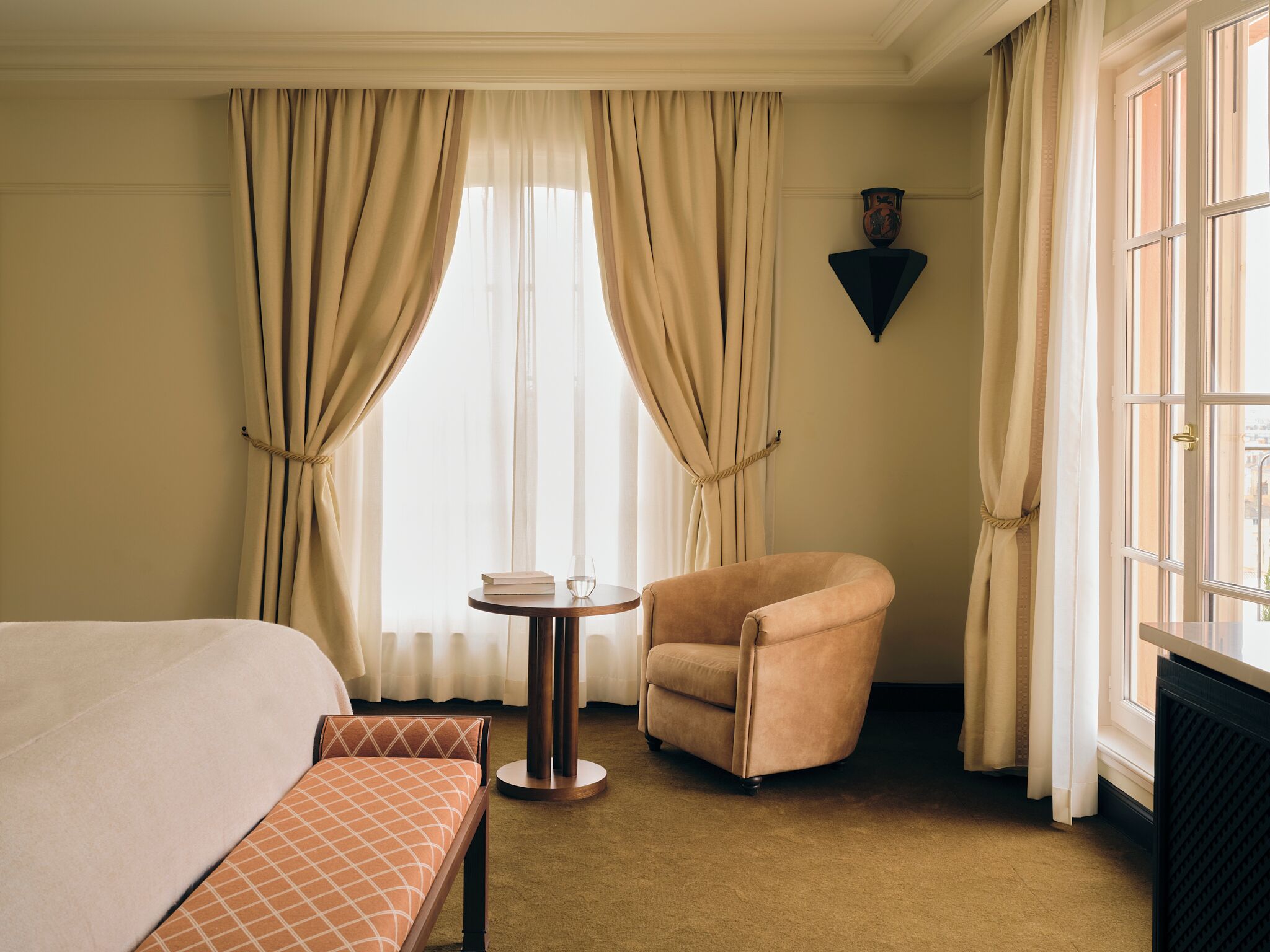Una habitación de hotel con cama, una silla, una mesita y ventana con cortinas.