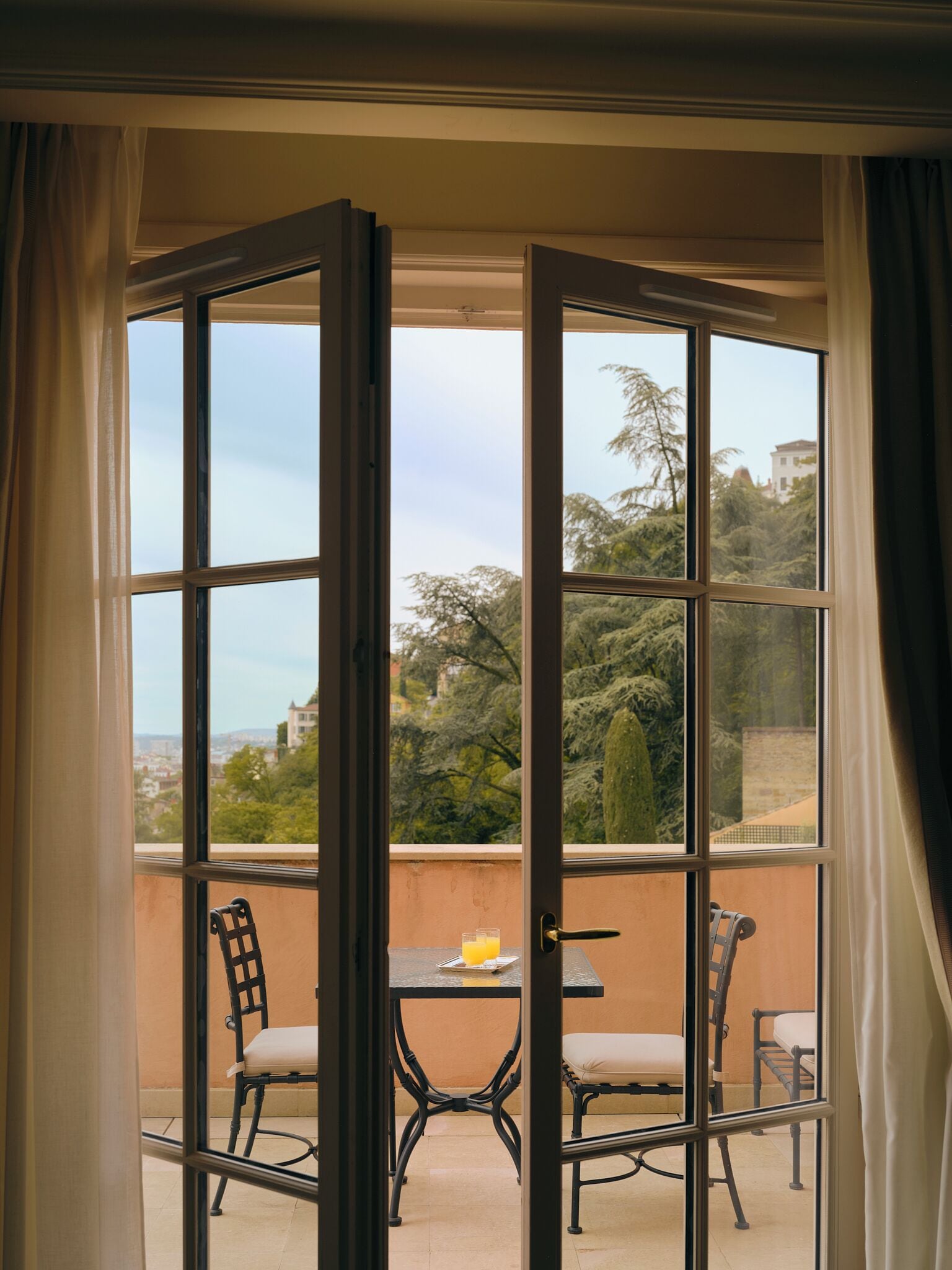 Quarto com uma janela grande, uma mesa com cadeiras e vista para áreas verdes e edifícios.