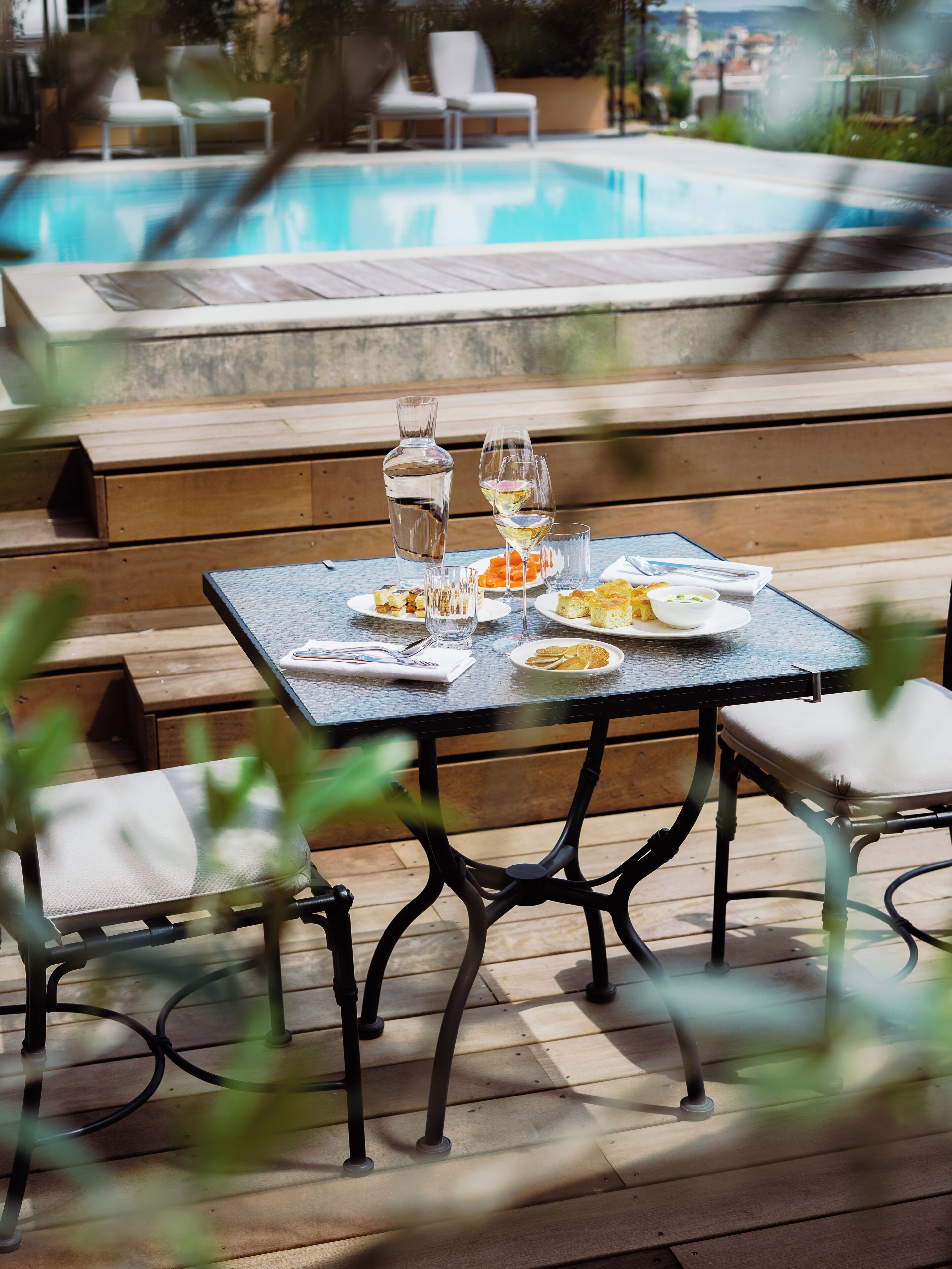 A dining table with plates, glasses, and a bottle, set on a wooden deck with a pool in the background.