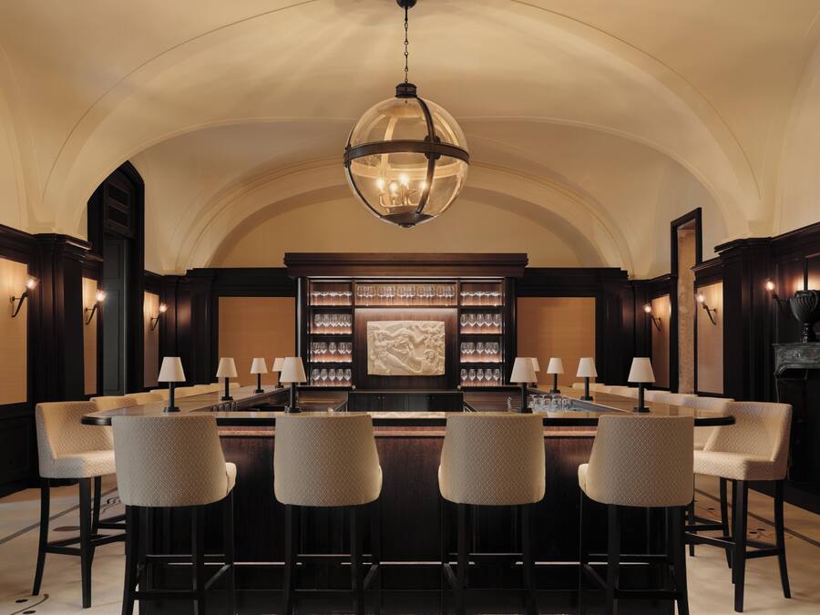 A bar area with a high ceiling and a central chandelier, surrounded by bar stools and a display of glasses.