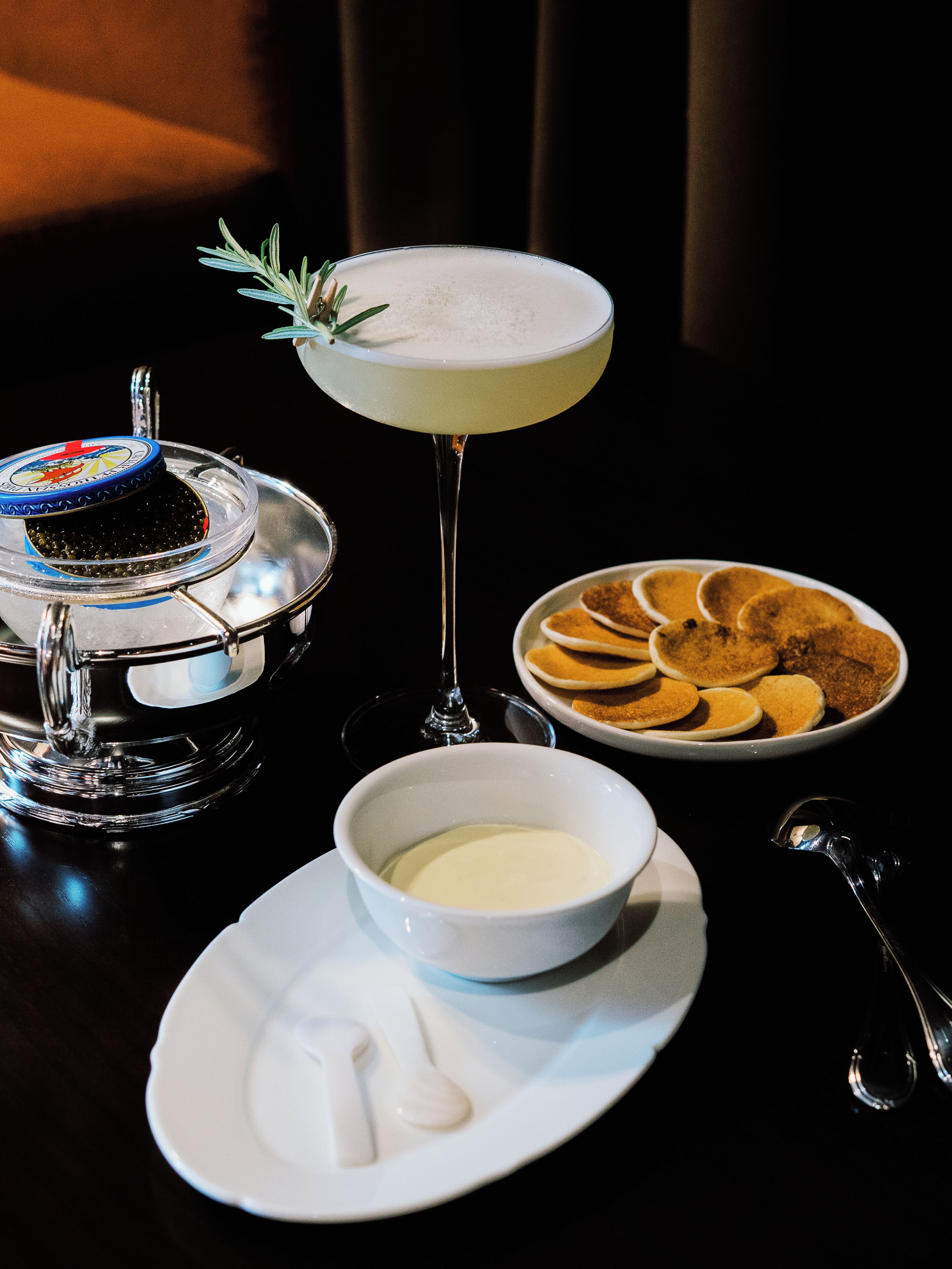 A table setting with a martini glass, a bowl of soup, a plate of pancakes, and a container of caviar.