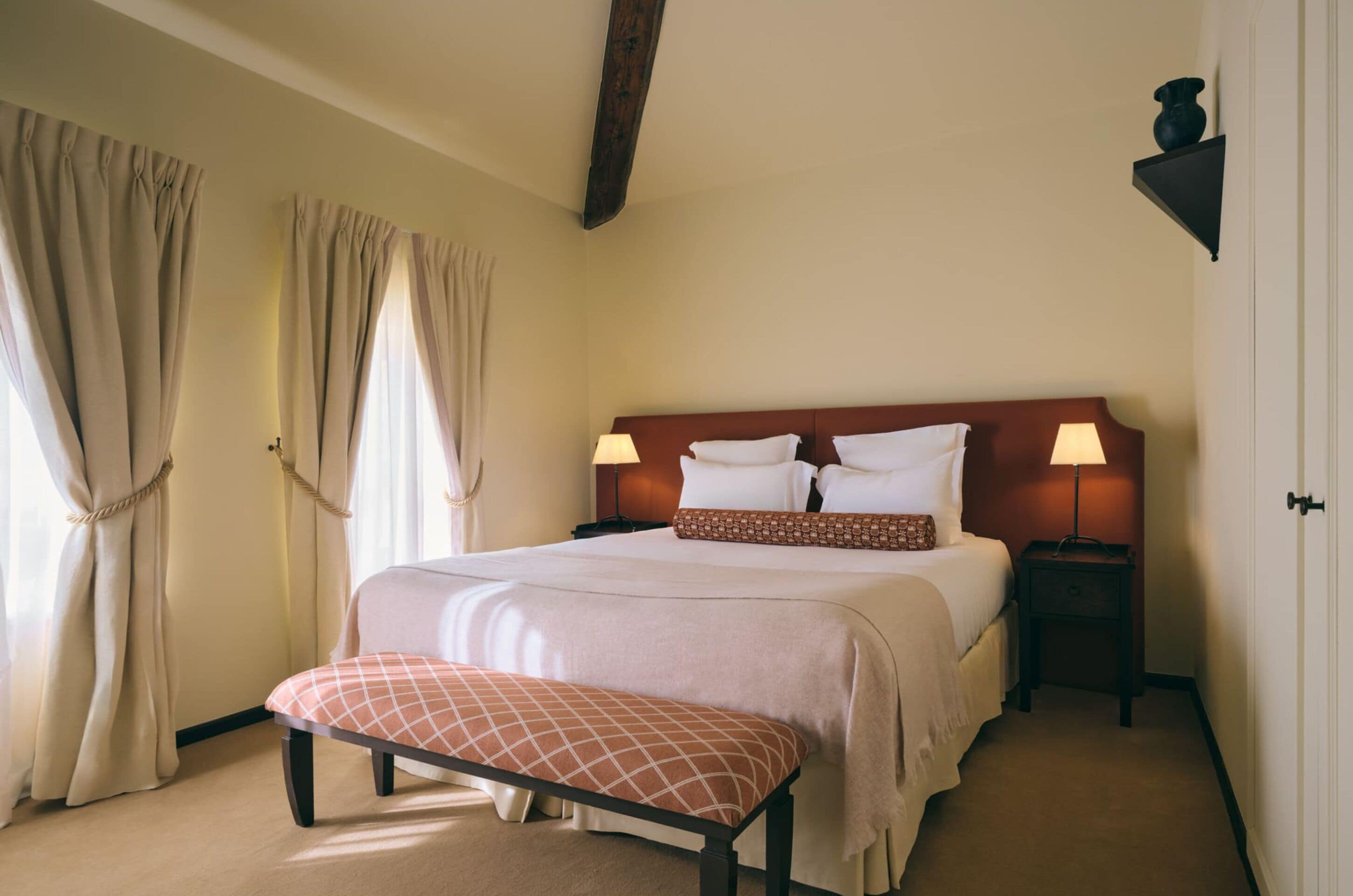 Habitación de hotel con cama, mesitas de noche, un banco, cortinas y una viga de techo de madera.