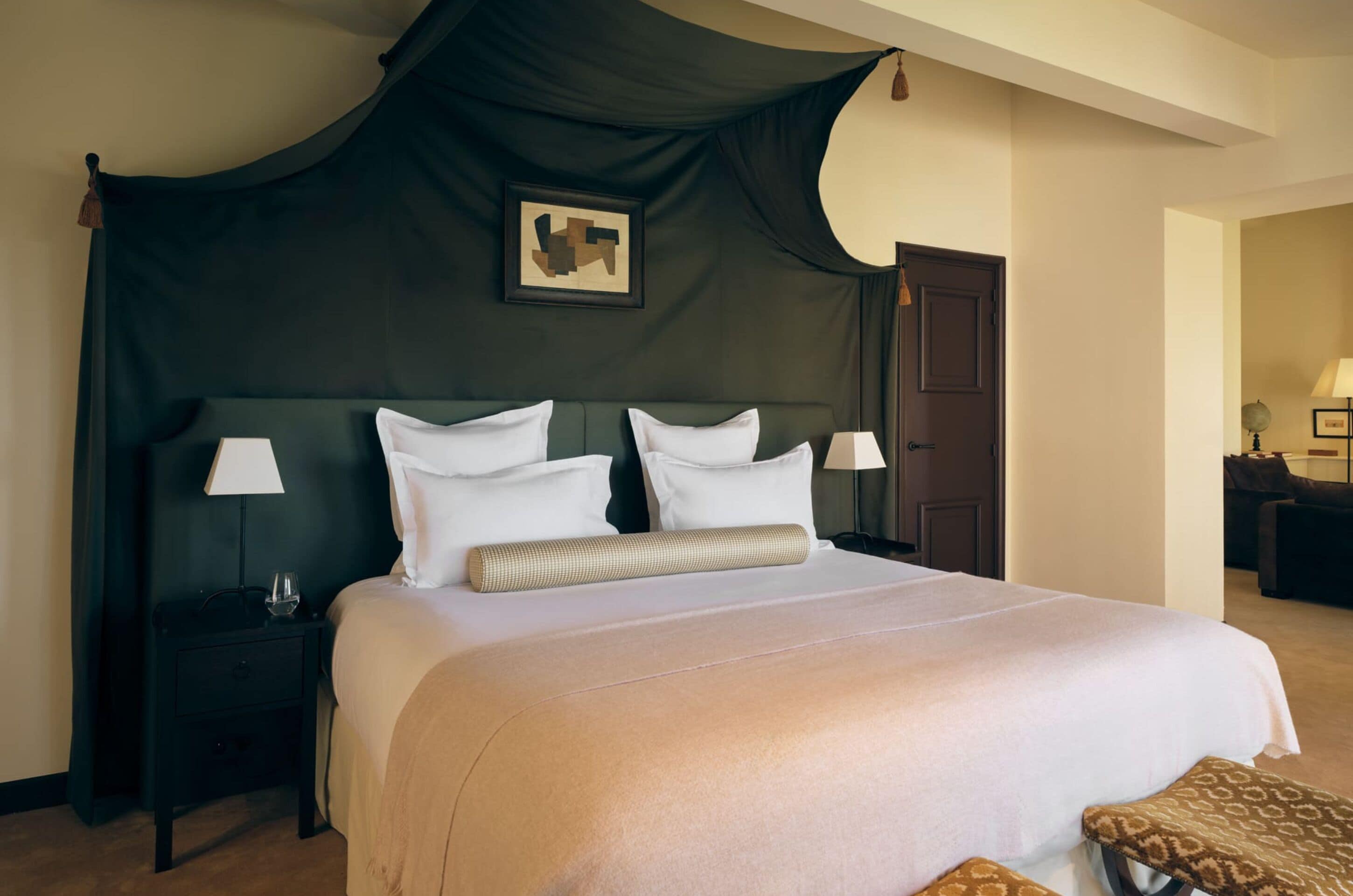 Habitación de hotel con una cama grande, dos mesitas de noche, una colcha oscura con dosel y un cuadro en la pared.