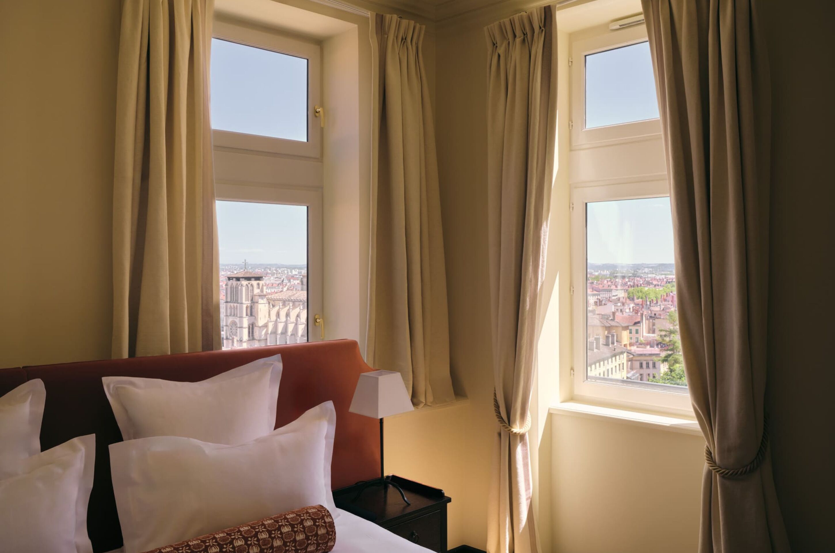 A hotel room with a bed, two windows with curtains, and a view of a cityscape.