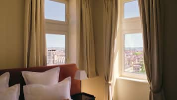 A hotel room with a bed, two windows with curtains, and a view of a cityscape.