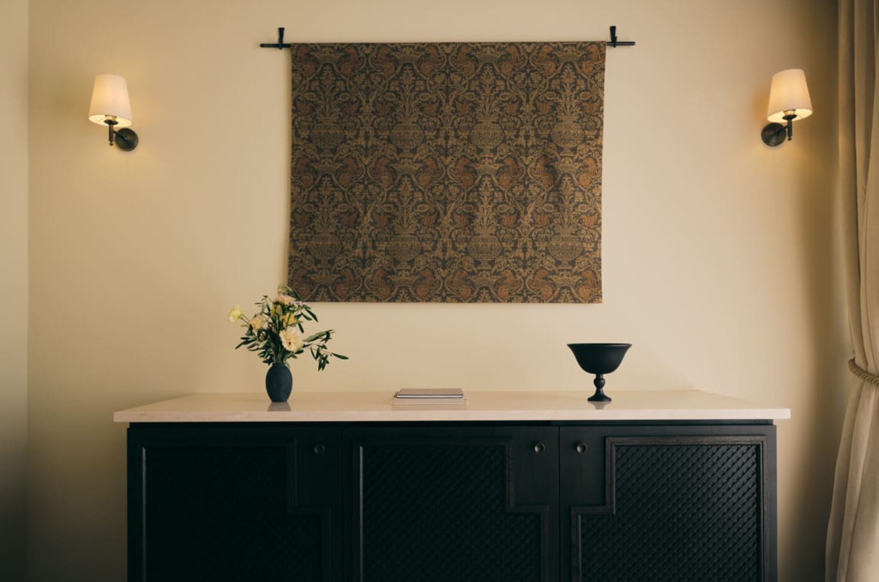 A room with a patterned tapestry hanging on the wall, a vase with flowers on a cabinet, and two wall-mounted lamps.