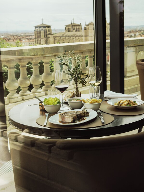 Una mesa de comedor con diversos platos, copas y un jarrón de flores, frente a una vista panorámica de la ciudad.