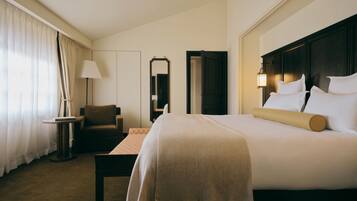 Quarto de hotel com uma cama grande, uma mesa de cabeceira, uma cadeira, um abajur, um espelho e uma porta.
