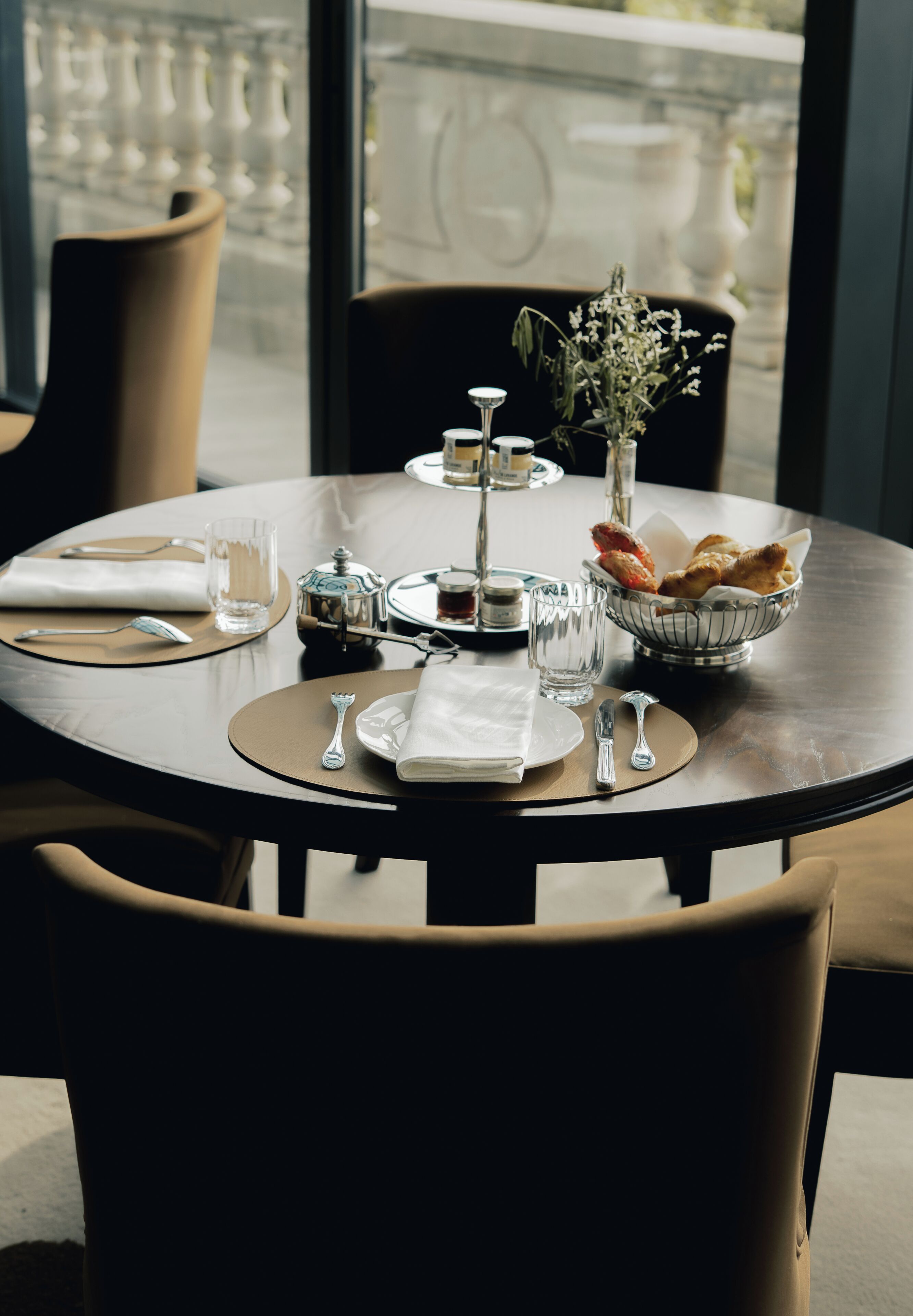 A round dining table set with plates, cutlery, glasses, a teapot, a vase with flowers, and a bowl of bread.