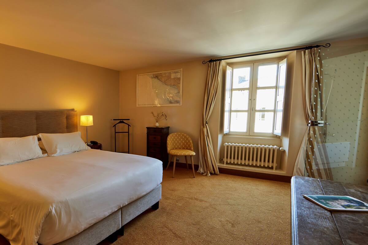 A hotel room with a large bed, a bedside table, a lamp, a chair, a radiator, and a window with curtains.