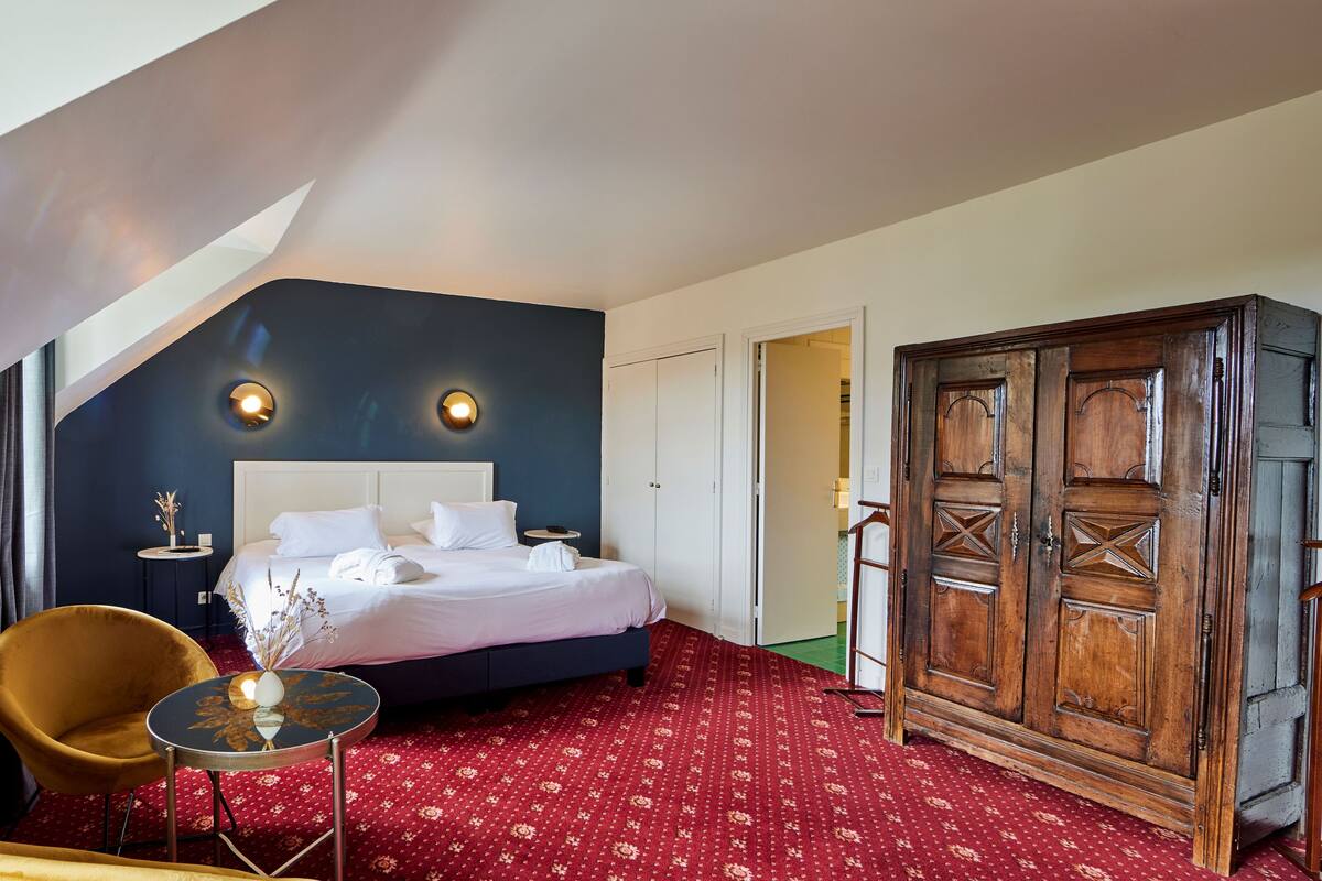A hotel room with a bed, a wooden wardrobe, a round table with a vase, a chair, and a small table with a lamp.