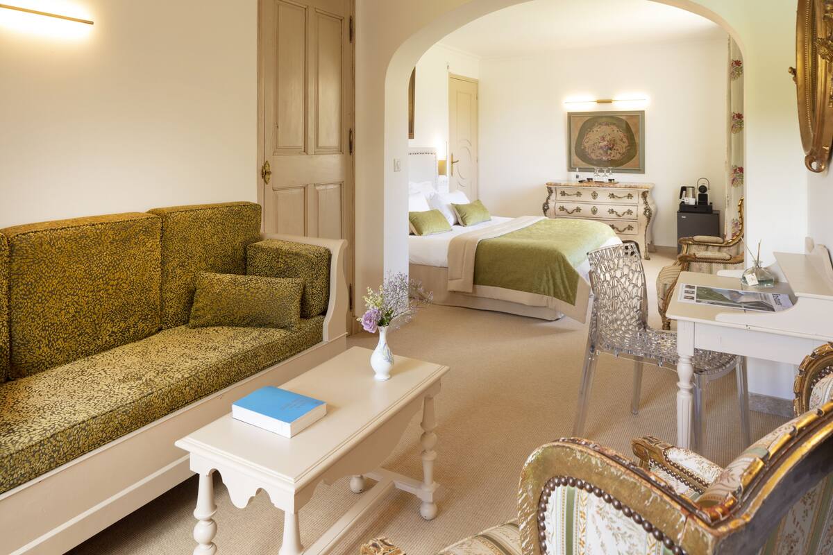 Una habitación con un sofá verde estampado, una mesita de centro blanca con un libro, una cama con ropa de cama verde, un aparador blanco y un espejo.