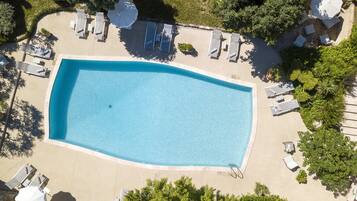 An aerial view of a rectangular swimming pool surrounded by lounge chairs and umbrellas, with trees and a building in the background.