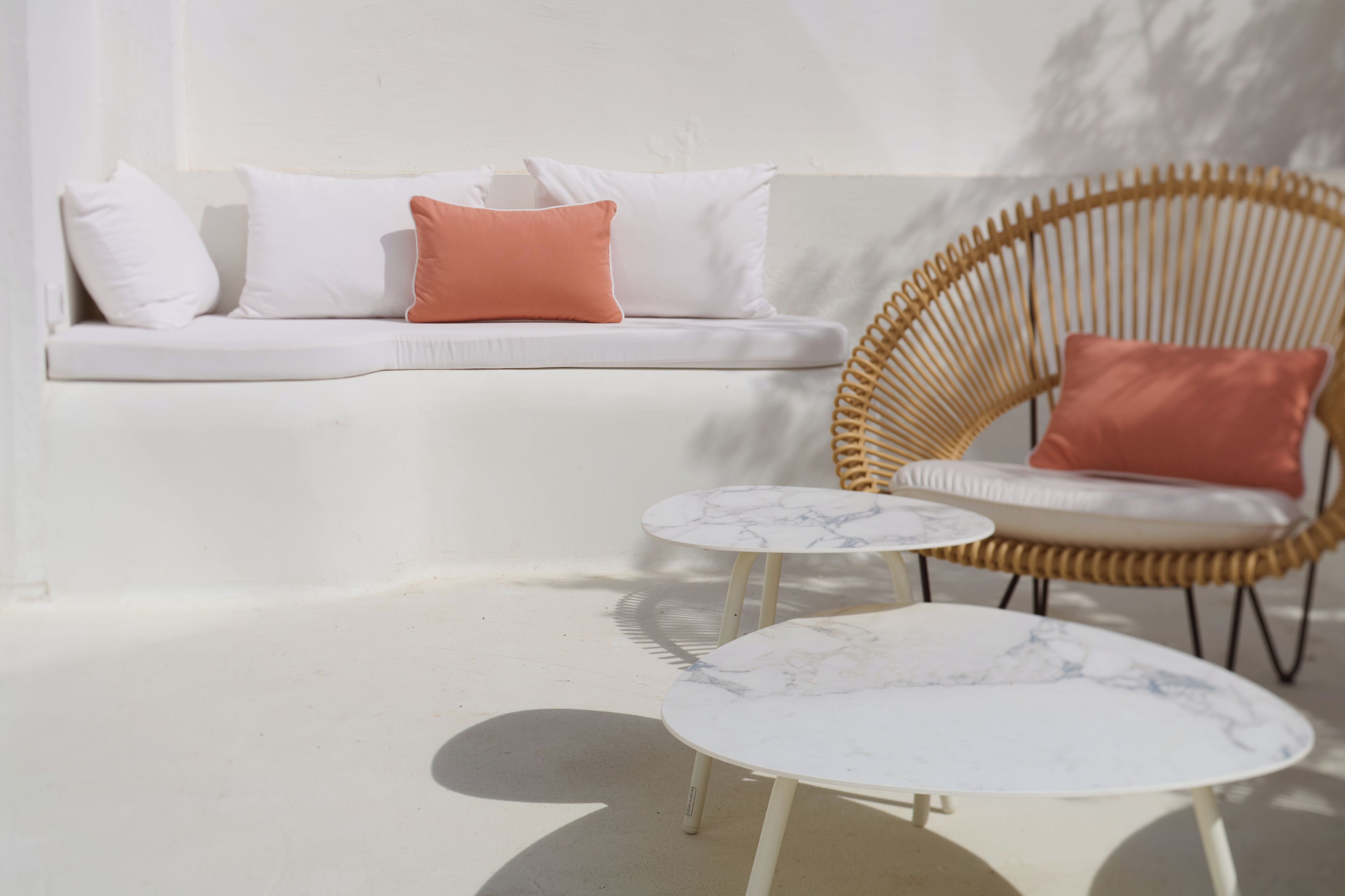 A white sofa with cushions, a wicker chair, and two marble tables.