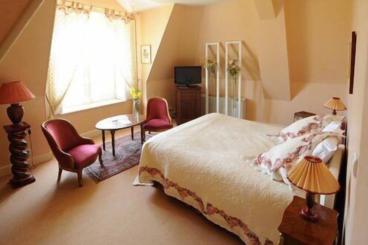 A bedroom with a bed, two chairs, a small table, a TV, and a window with curtains.