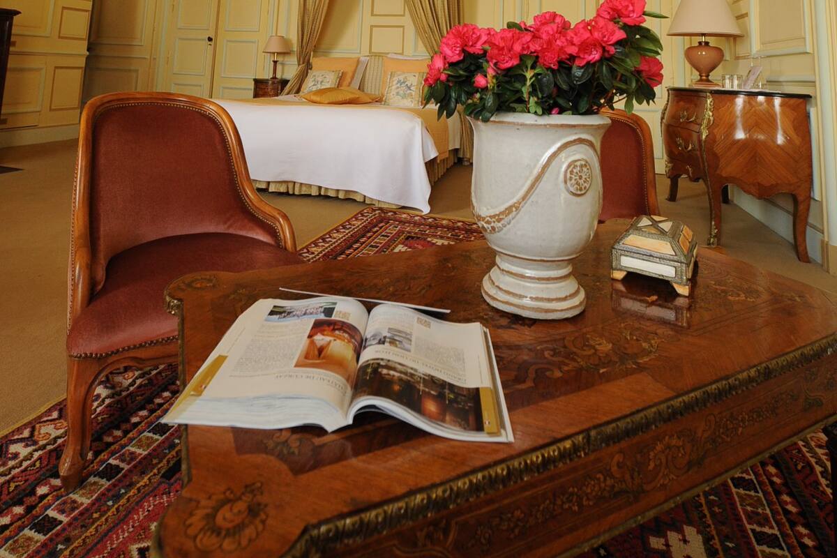 A hotel room with a bed, a red armchair, a wooden coffee table with a magazine, a vase with flowers, and a lamp.