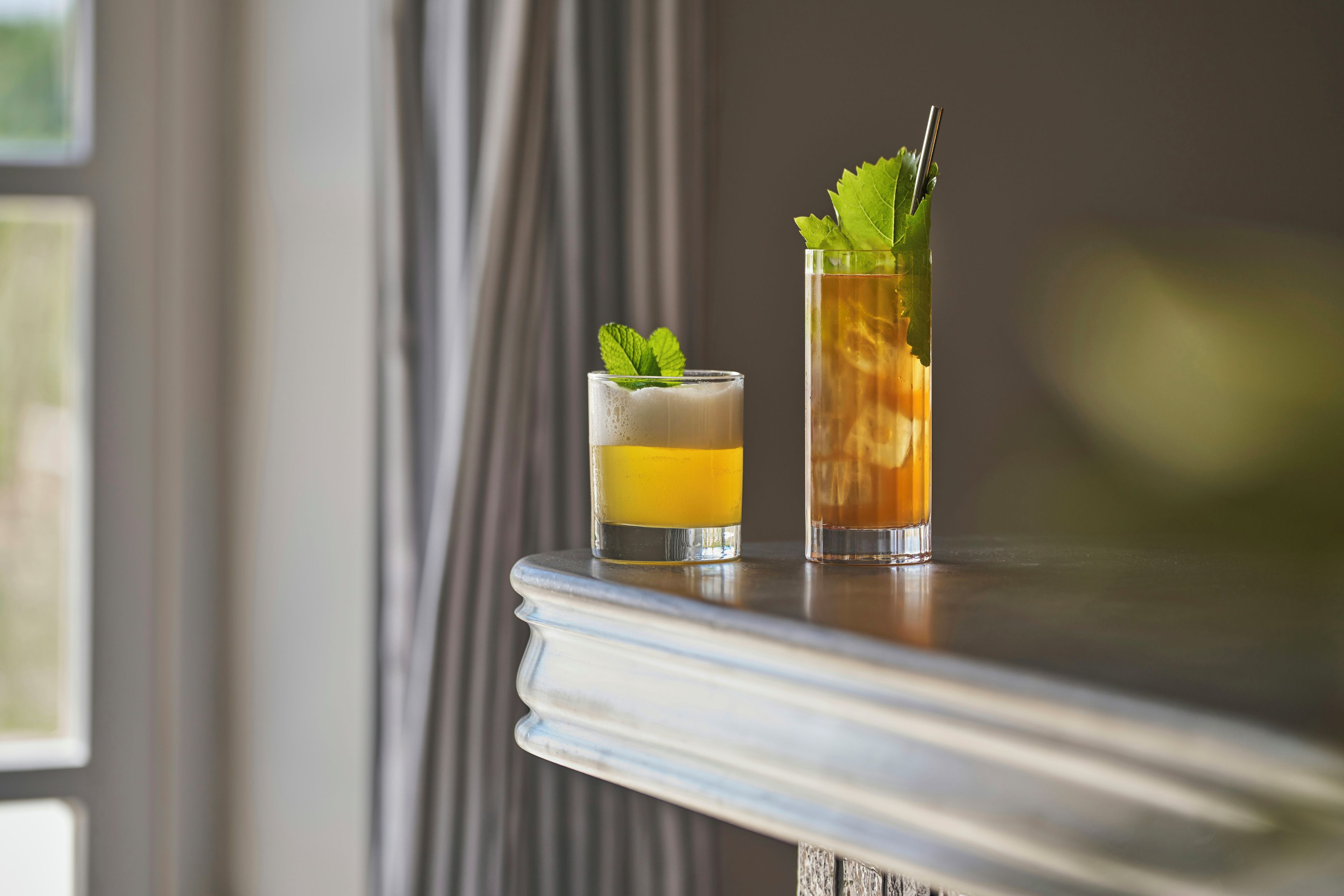 Two glasses of beverages on a table with a window in the background.