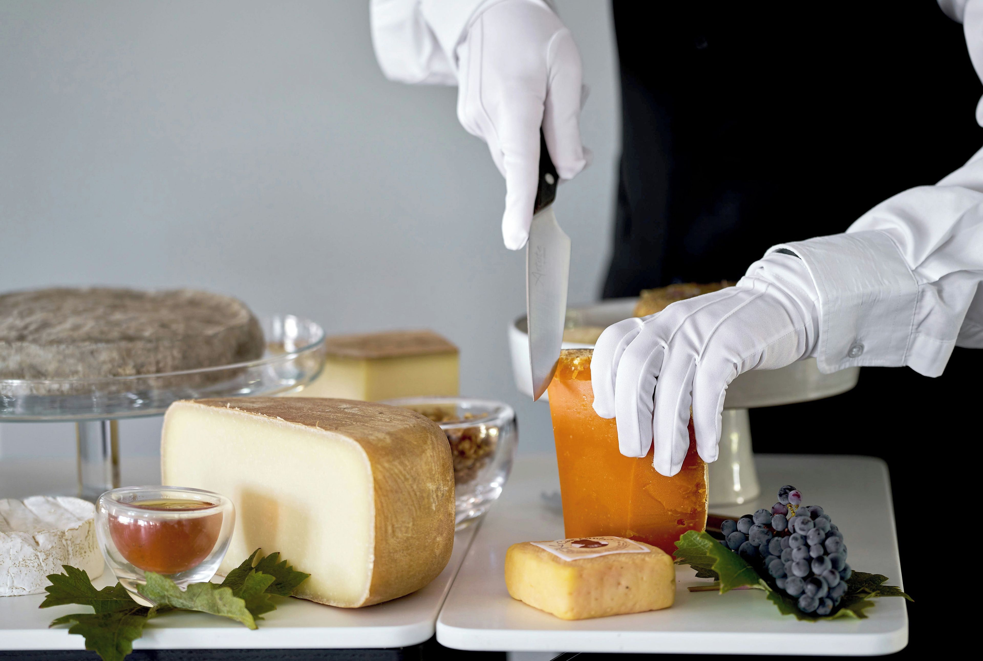 A person cutting a wheel of cheese with a knife, surrounded by various cheeses and a bowl of honey.