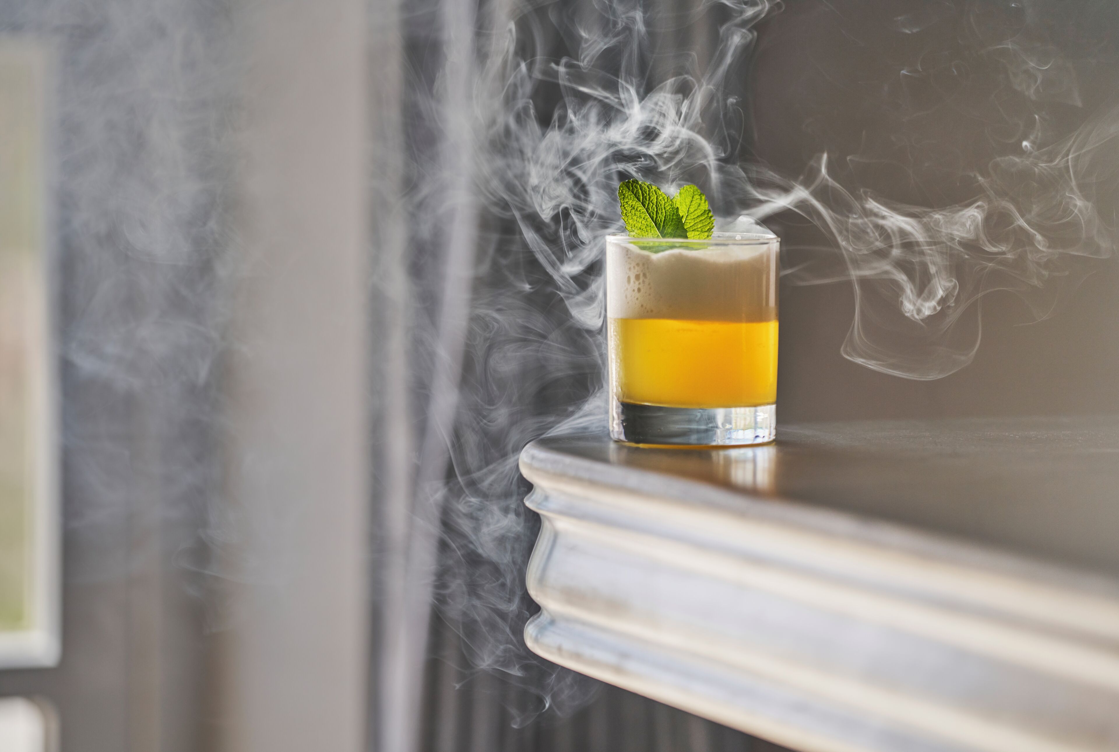 Un vaso con una bebida en capas y una ramita de menta, rodeado de humo, sobre una superficie reflectante.