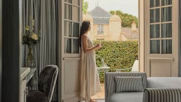 A woman standing by a window with a view of a building and a hedge.
