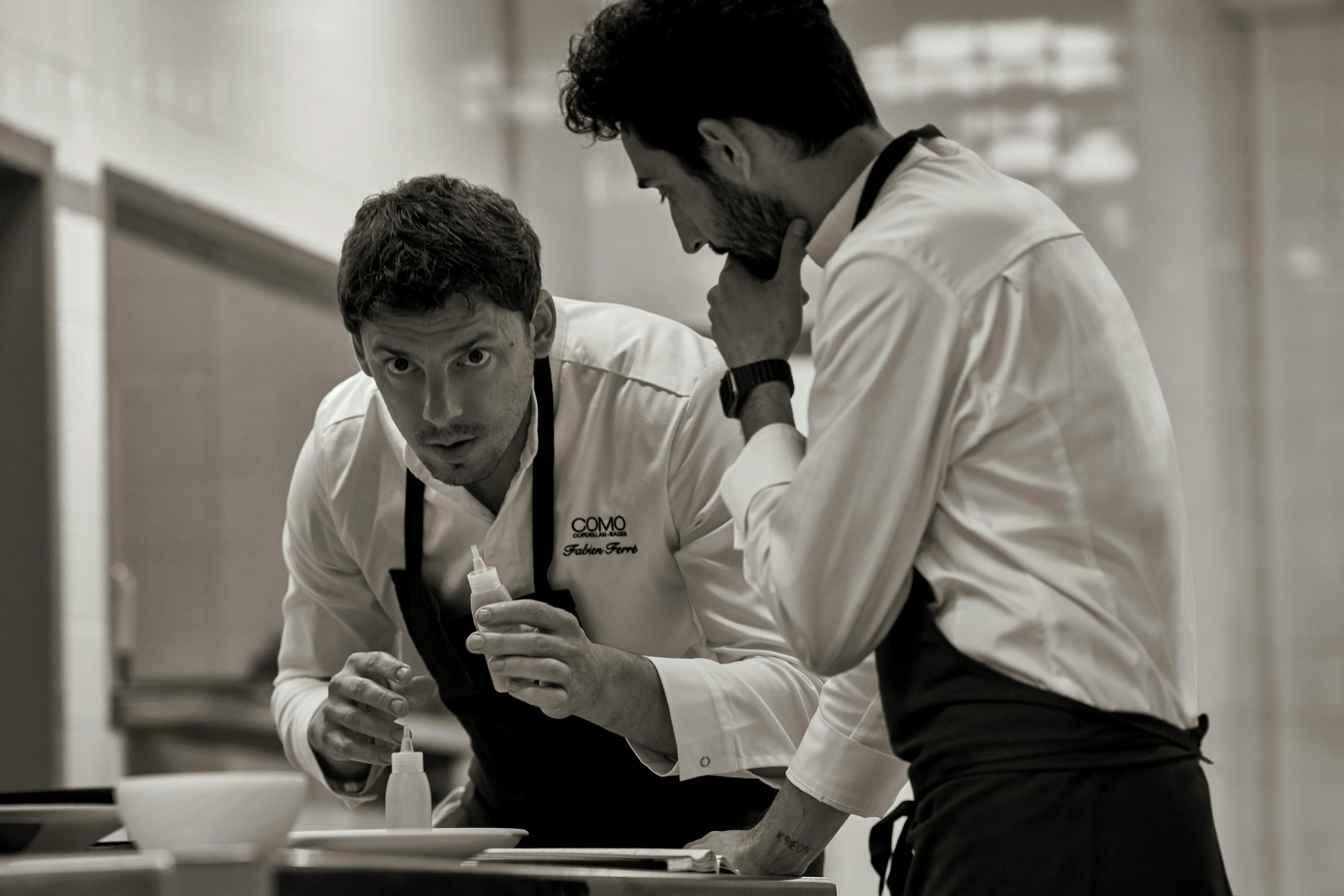 Two individuals in a professional setting, possibly a kitchen, with one person holding a bottle.