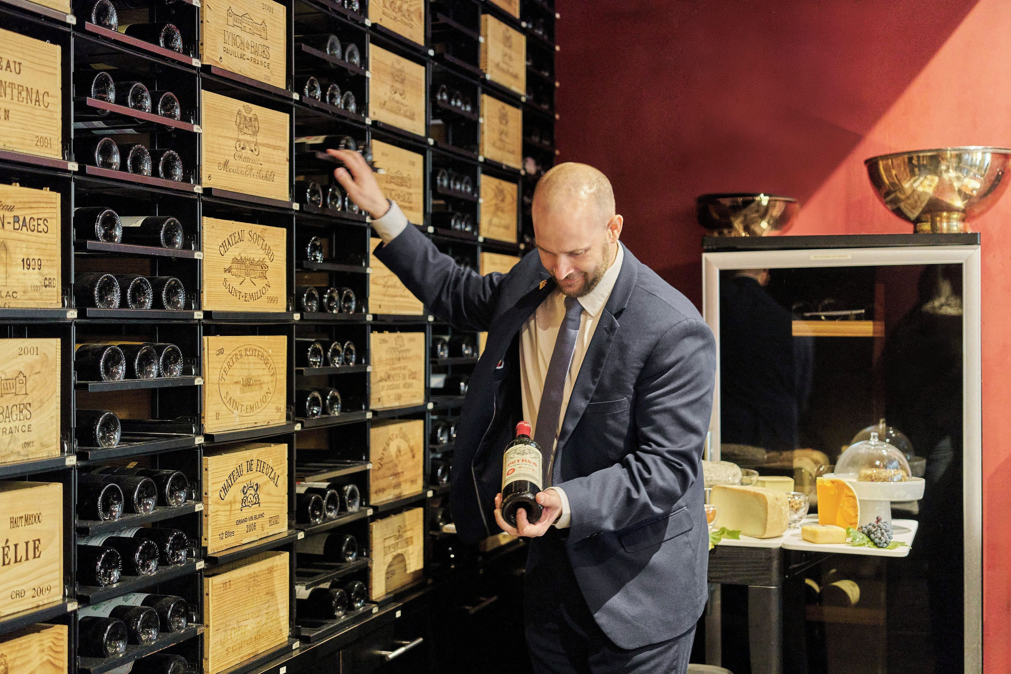 A man in a suit examining wine bottles in a wine cellar with wooden racks and labels.