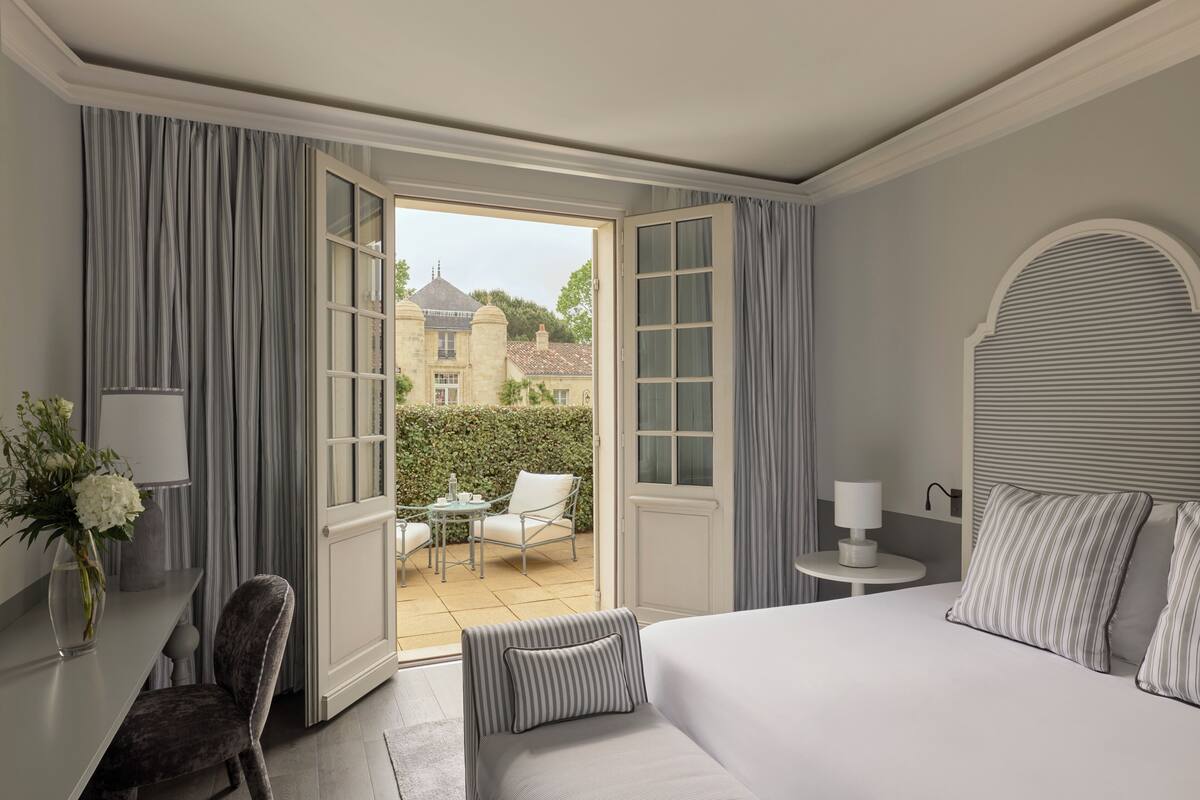 A hotel room with a bed, a desk, a chair, a small table, and a view of the outside.