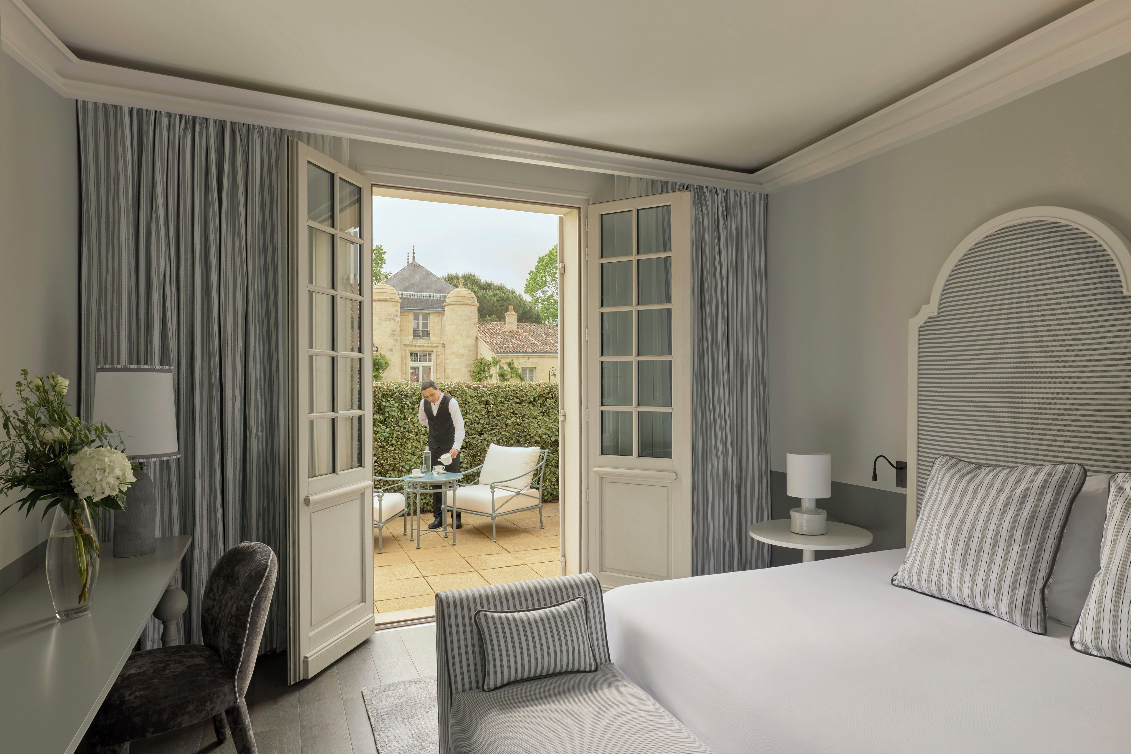 A hotel room with a bed, a desk, a chair, a small table, and a view of a courtyard with a person outside.