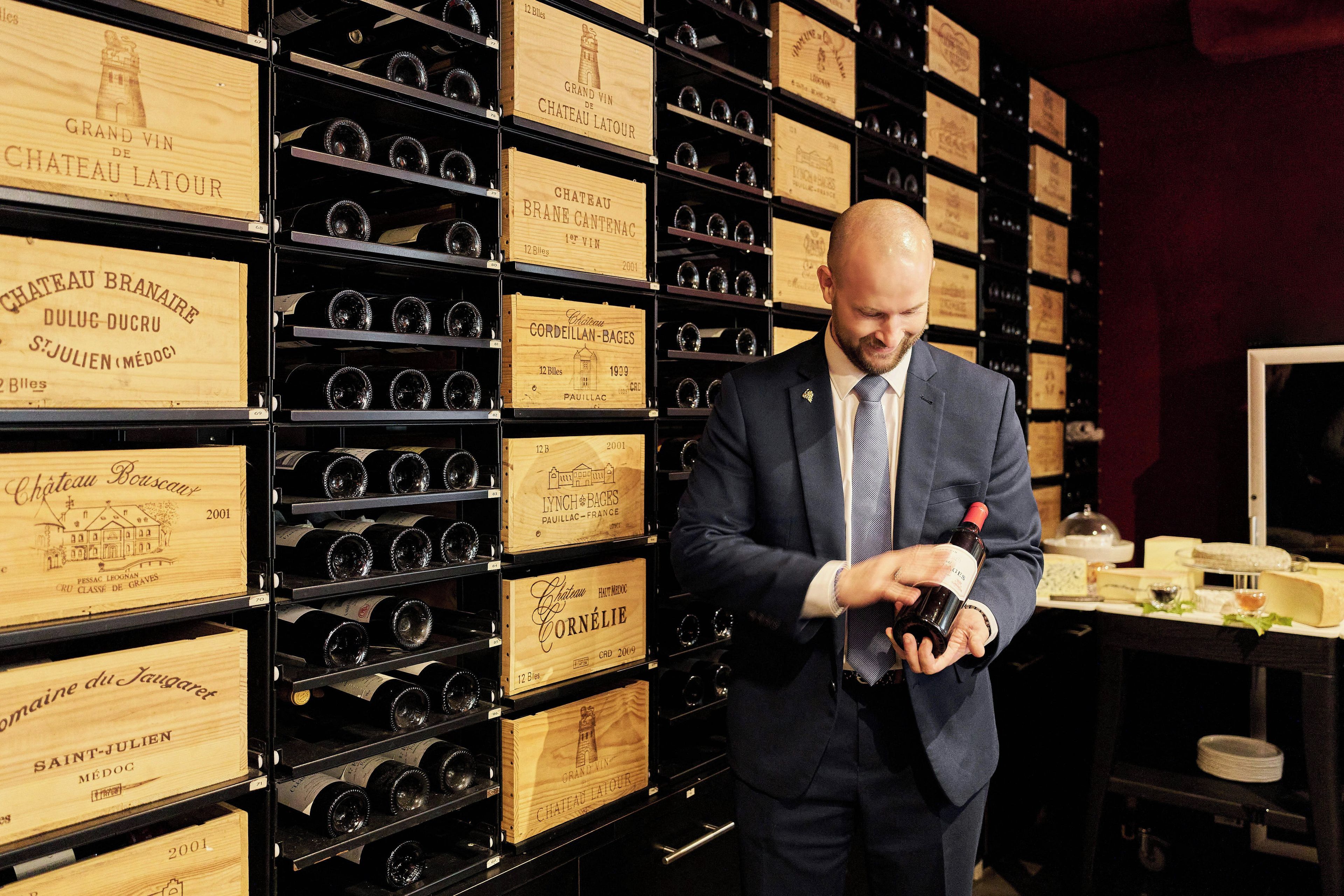 A man in a suit holding a bottle of wine in front of a wall of wine bottles in wooden boxes.