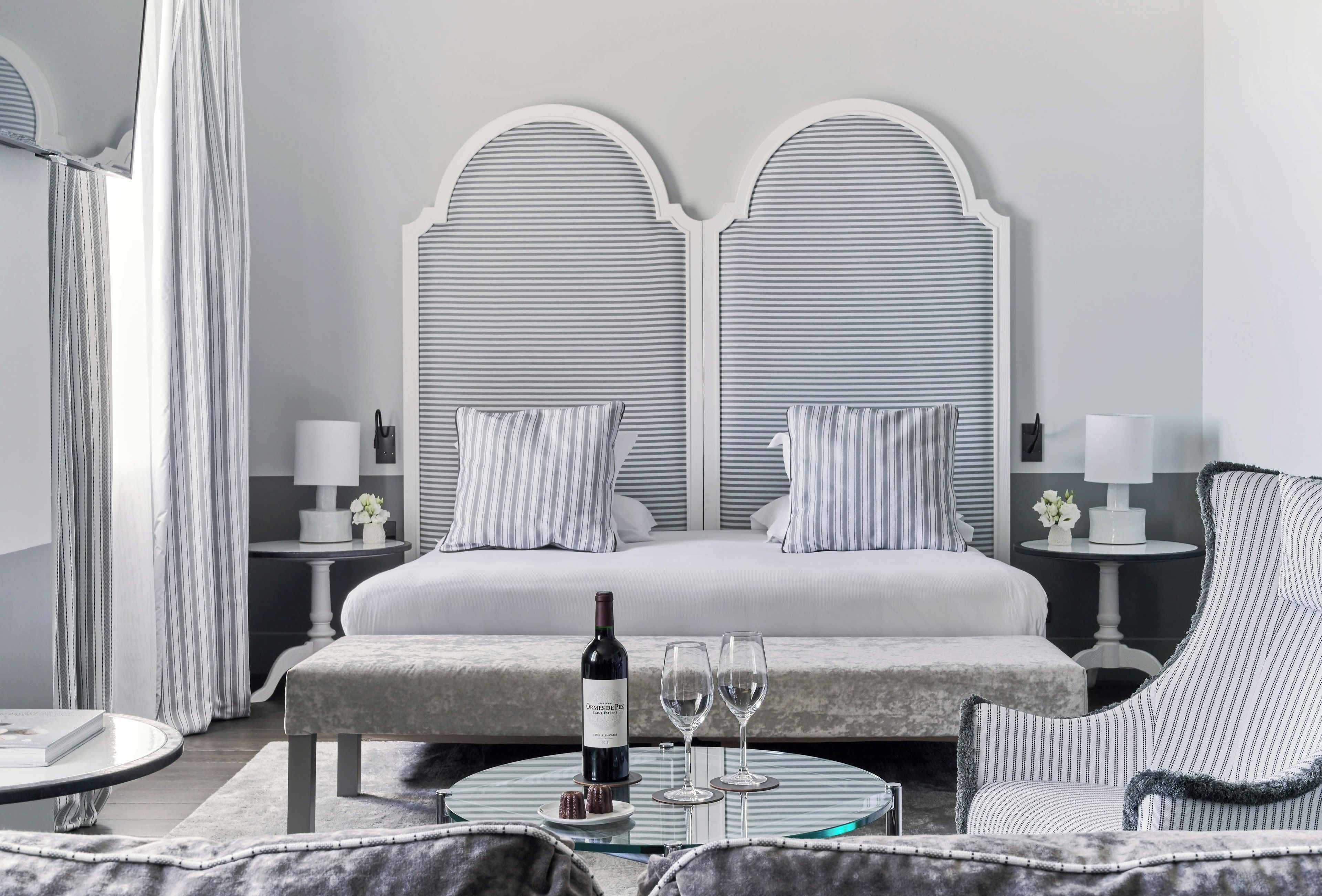 A hotel room with a large bed, two bedside tables, a round glass table with a bottle and glasses, and a chair.
