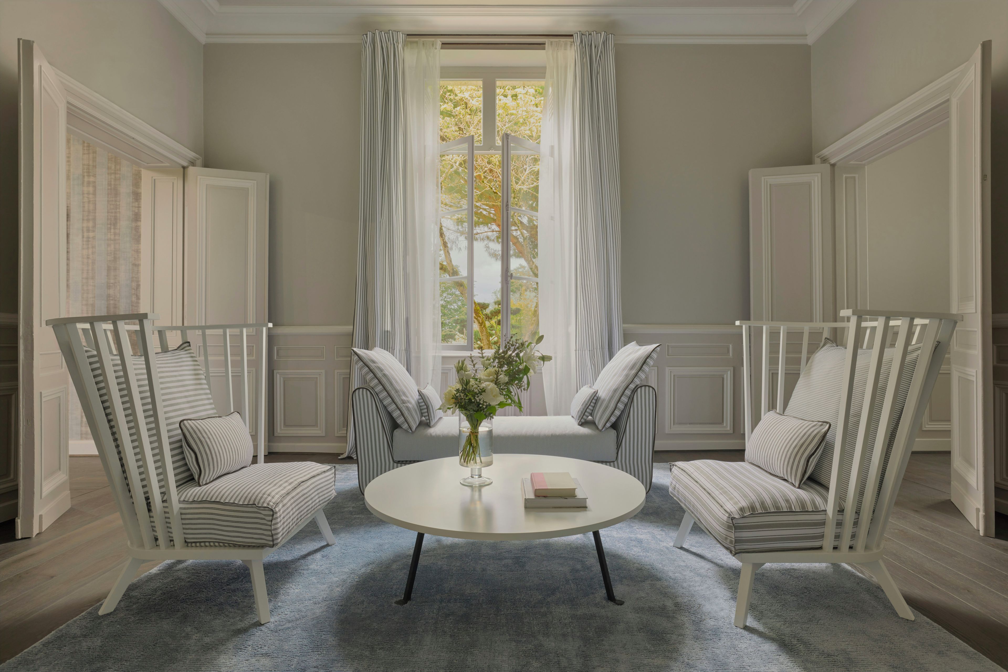 Una habitación con una mesa redonda, dos sillones a rayas, un sofá y un ventanal con cortinas.
