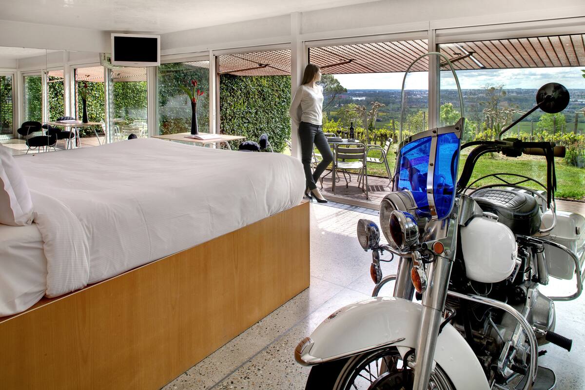 A bedroom with a large bed, a motorcycle, and a view of the countryside.
