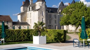 A swimming pool with lounge chairs and umbrellas, a stone building with multiple towers in the background, and a clear blue sky. - Château de Marçay (Marcay)