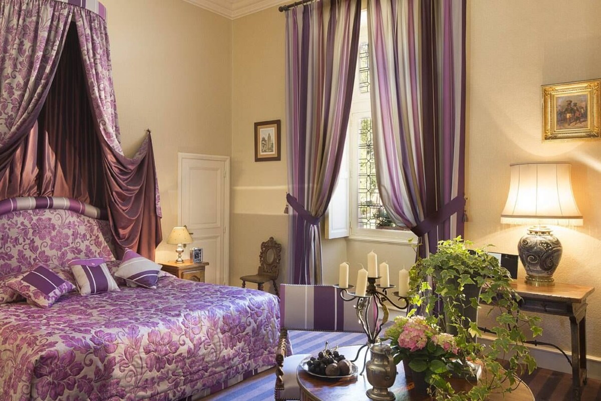 A bedroom with a four-poster bed, a bedside table with a lamp, a vase with flowers, and a window with curtains.