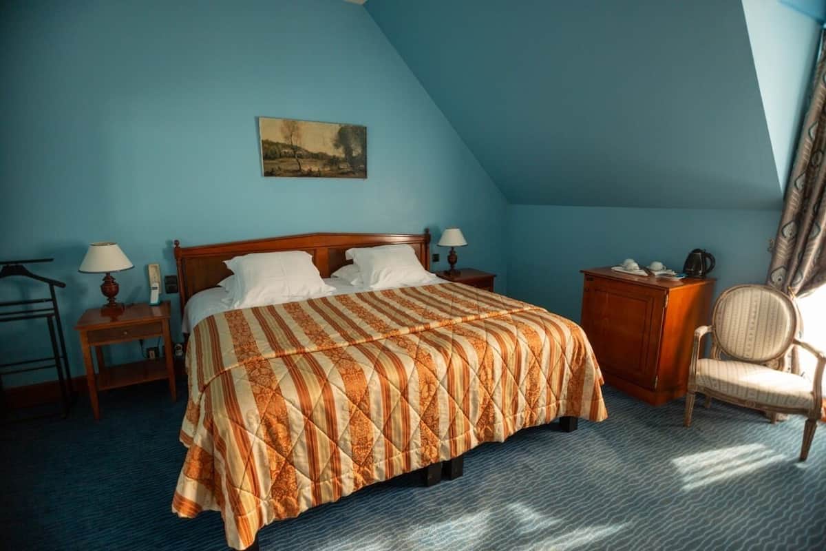 Un dormitorio con una cama, dos mesitas de noche, una silla, una mesita auxiliar y un cuadro en la pared.