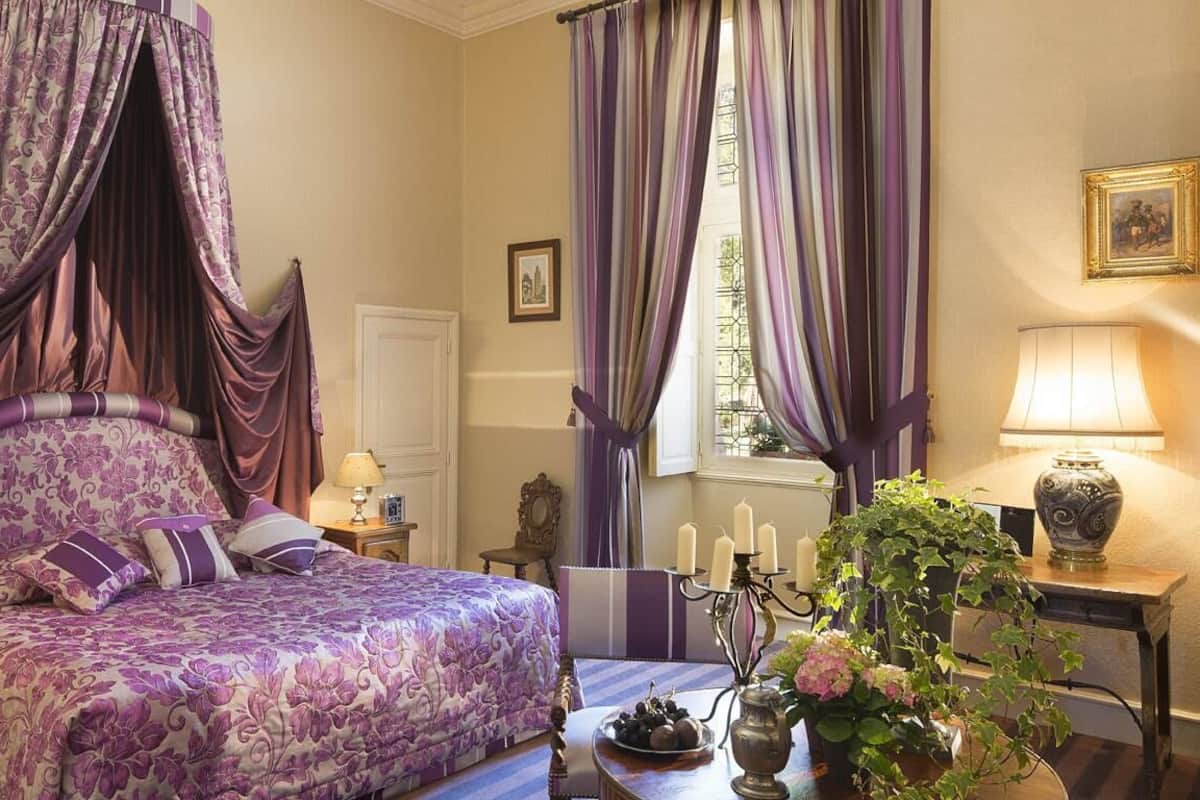 Un dormitorio con cama con dosel, una mesita de noche con lámpara, un florero con flores y una ventana con cortinas.