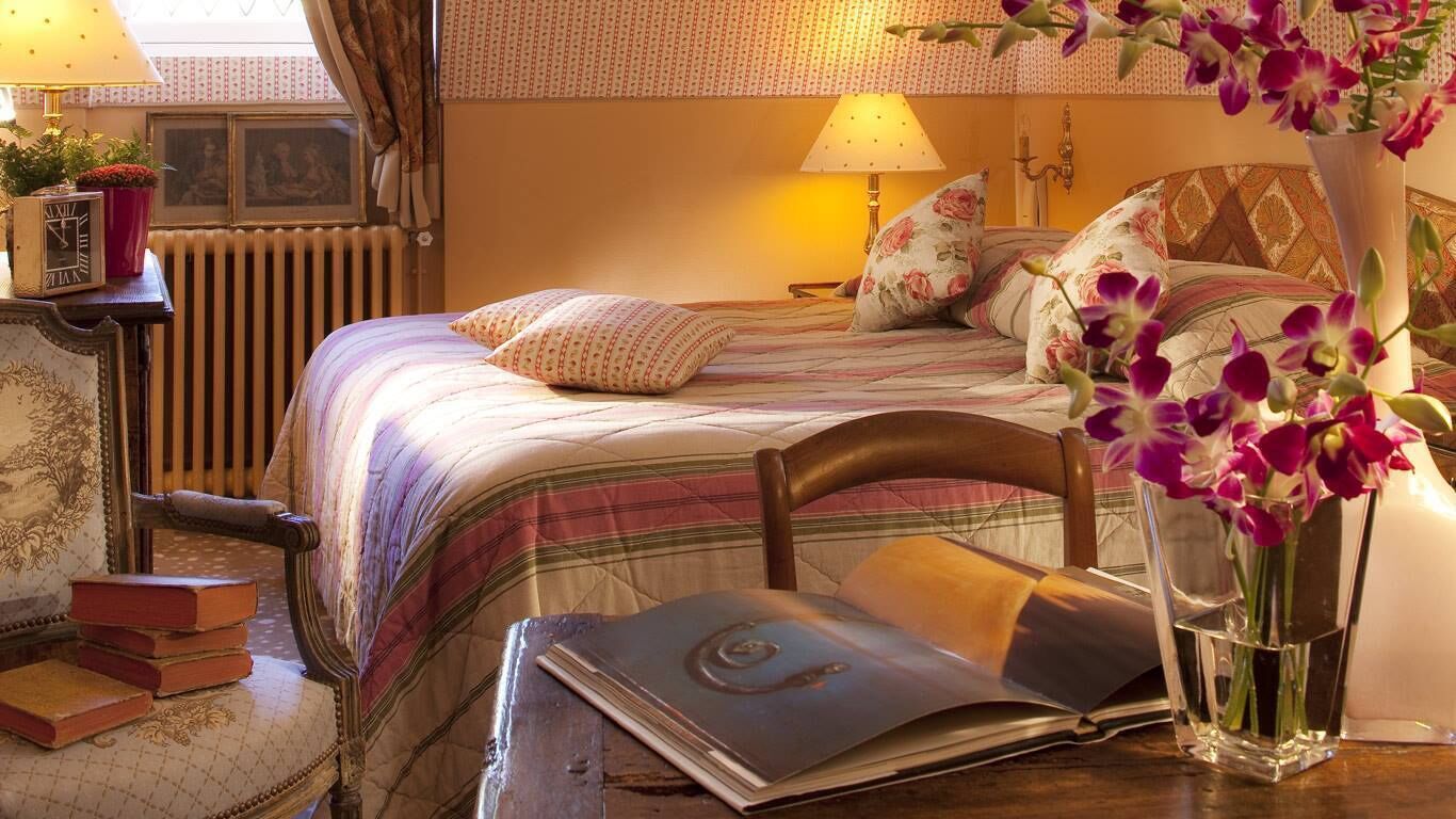 Une chambre à coucher bien rangée, avec un lit, des oreillers, une table de chevet avec une lampe, un livre et un vase rempli de fleurs.