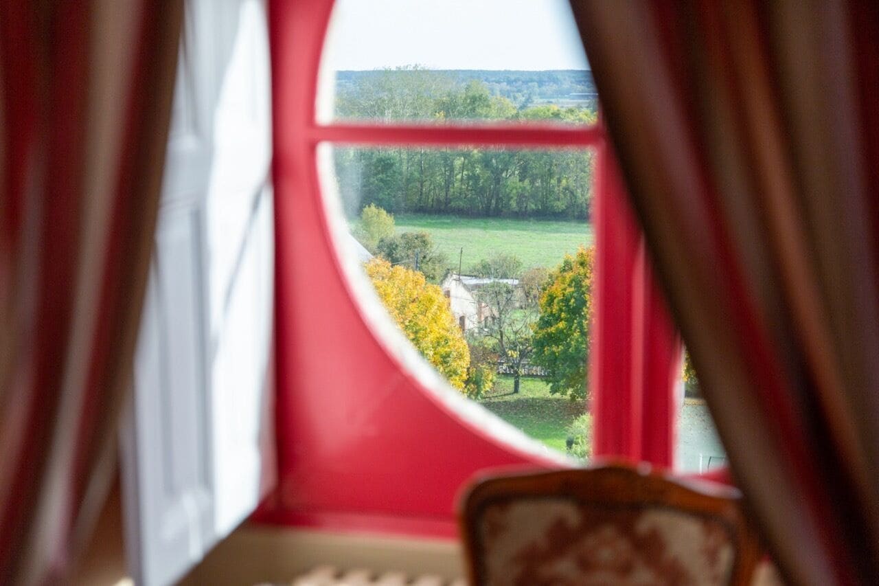 Una habitación con un ventanal grande, cortinas y vistas a un paisaje verde con árboles y un edificio.