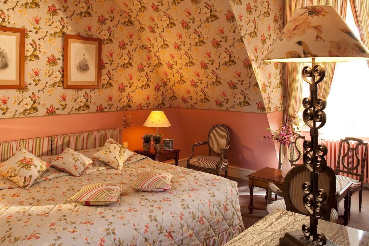 Un dormitorio con colcha de estampado floral, mesita de noche con lámpara, una silla y cuadros en la pared.