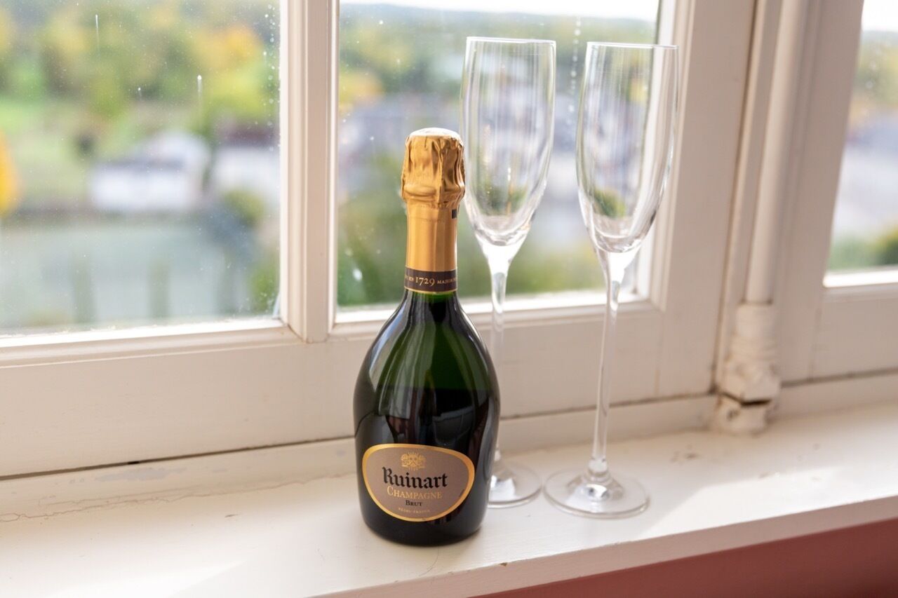 A Ruinart Champagne bottle and two glasses on a windowsill with a view of a body of water and greenery.