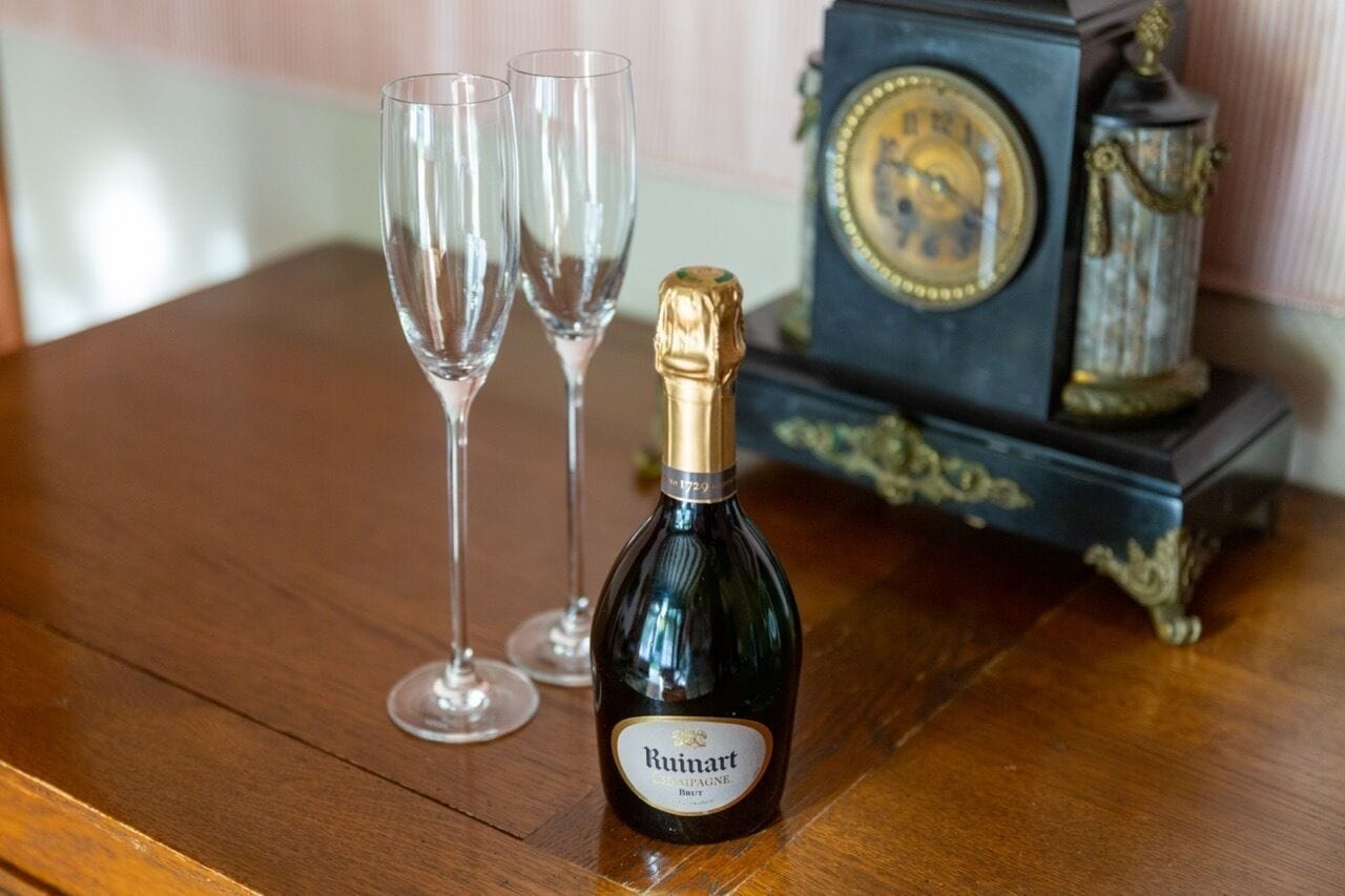 Una botella de champán Ruinart y dos copas vacías sobre una mesa de madera, con un reloj decorativo al fondo.