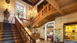 A grand staircase with ornate railings and a chandelier, leading to a high-ceilinged room with a wooden ceiling and a stained glass window.