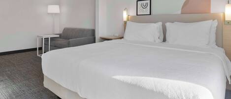 A hotel room with a large bed, a gray armchair, a small table, and a wall-mounted lamp.