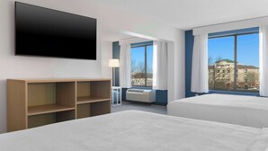 Une chambre d’hôtel avec deux lits, un téléviseur à écran plat, une étagère en bois, un bureau et une fenêtre donnant sur la ville.