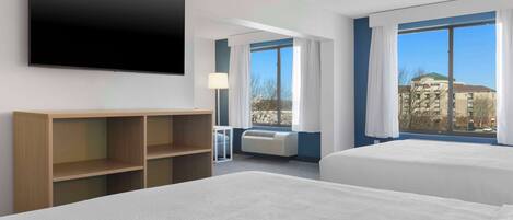 A hotel room with two beds, a flat-screen TV, a wooden shelf, a desk, and a window with a city view.