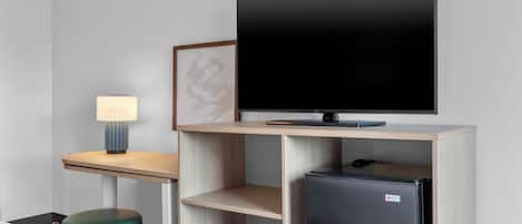 A modern hotel room with a flat-screen TV mounted on a wooden shelf, a small refrigerator, a green ottoman, and a desk with a lamp.