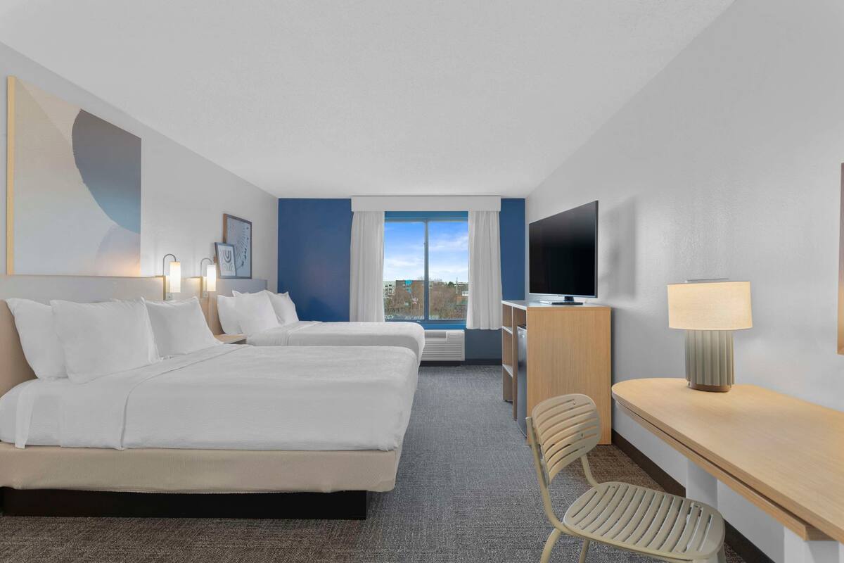 Une chambre d’hôtel avec deux lits, un bureau, une chaise, une télévision et une fenêtre avec des rideaux.